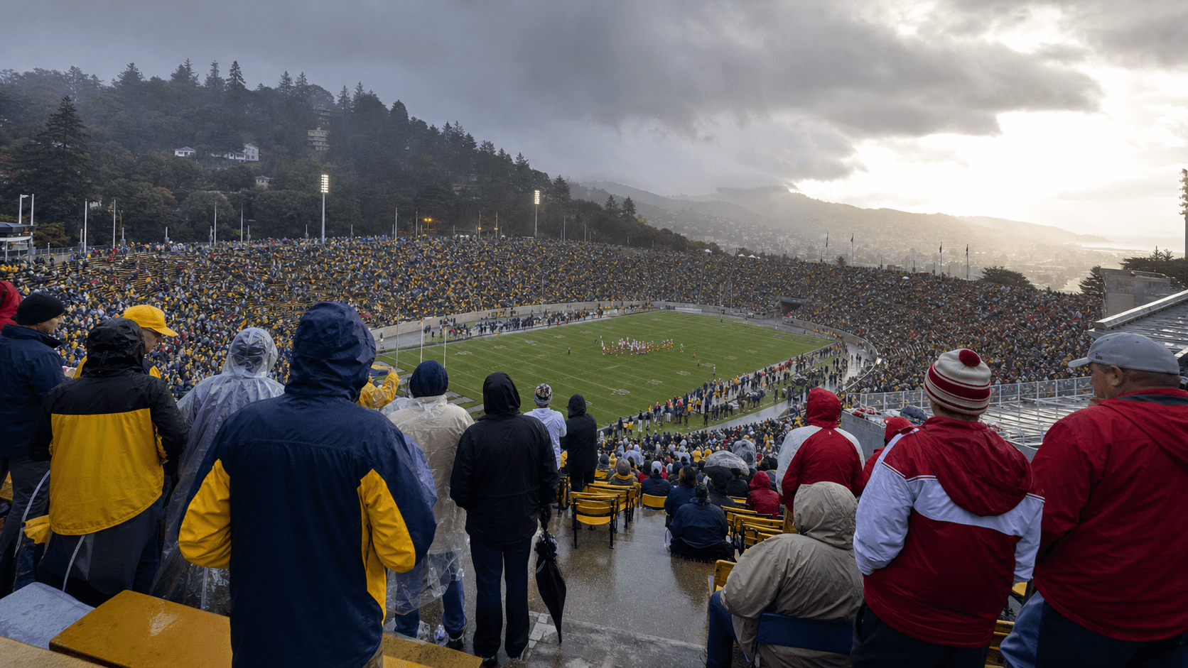 Big Game fans in rain gear watching a wet hillside stadium beneath clearing storm clouds