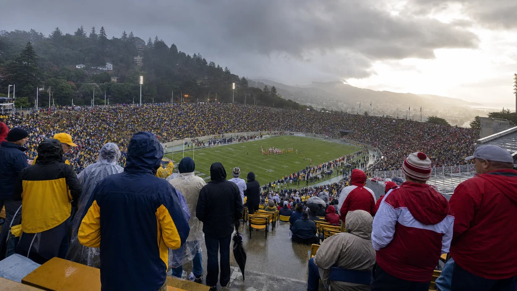 Big Game fans in rain gear watching a wet hillside stadium beneath clearing storm clouds