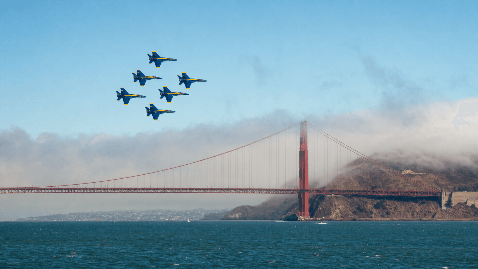 Six blue and gold demonstration jets flying over the Golden Gate Bridge as coastal fog enters San Francisco Bay
