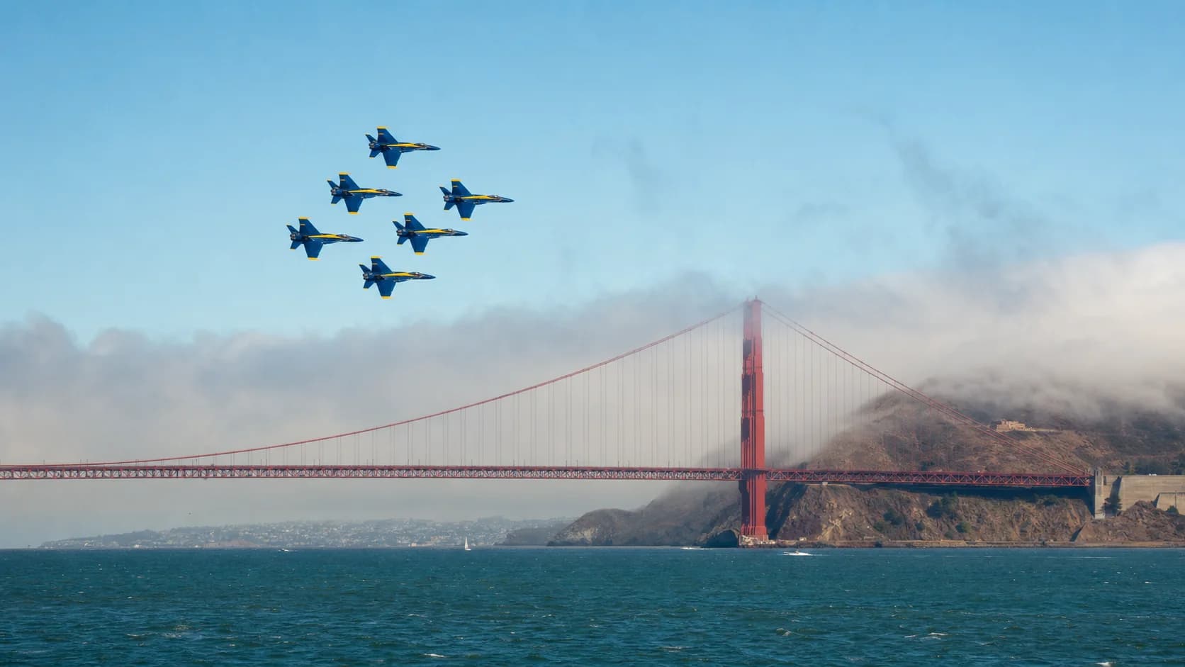 Six blue and gold demonstration jets flying over the Golden Gate Bridge as coastal fog enters San Francisco Bay