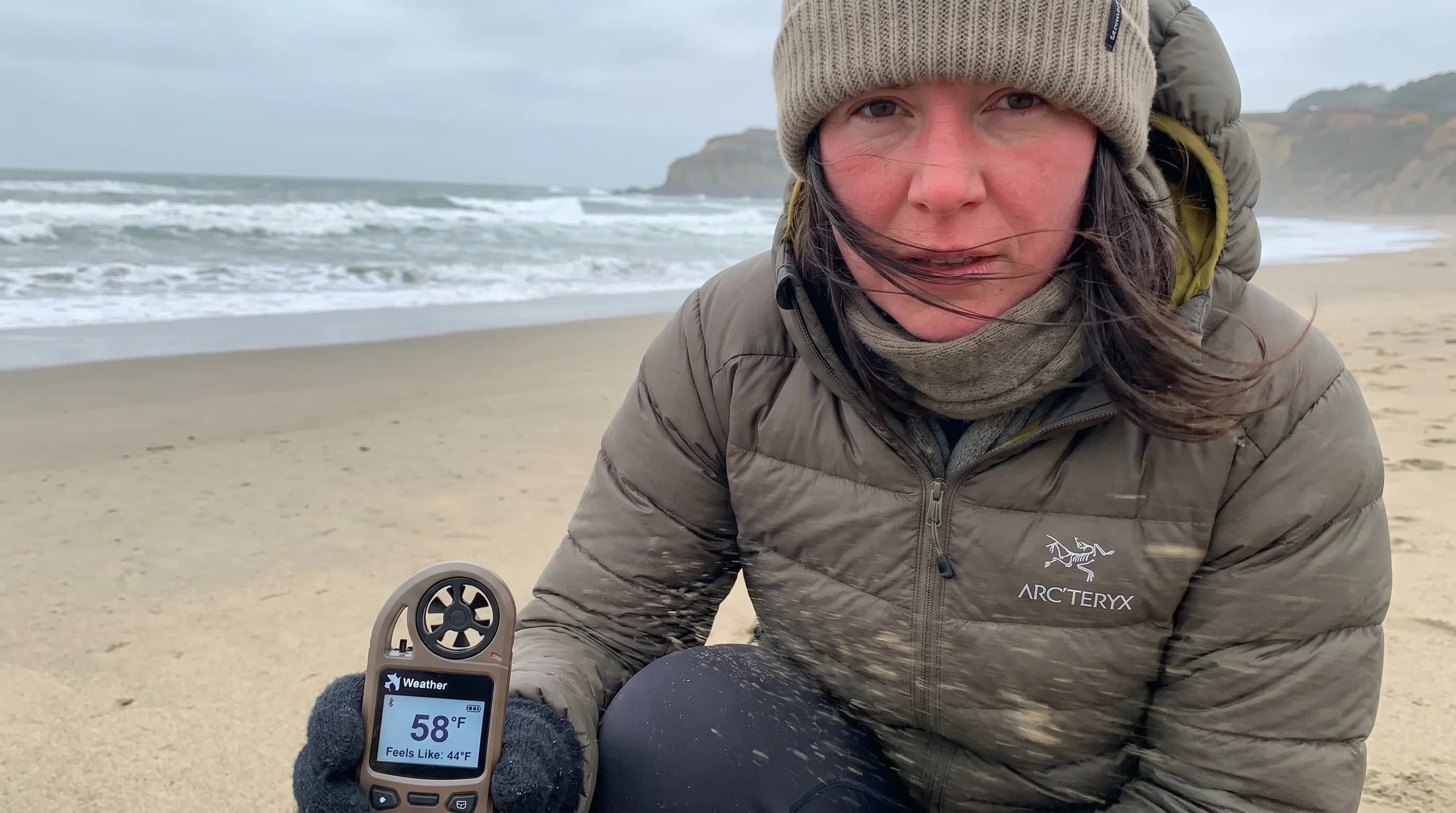 Person in jacket at breezy San Francisco coastal location, wind visibly moving hair, fog in background, thermometer showing 57°F with felt temperature noted as 44°F
