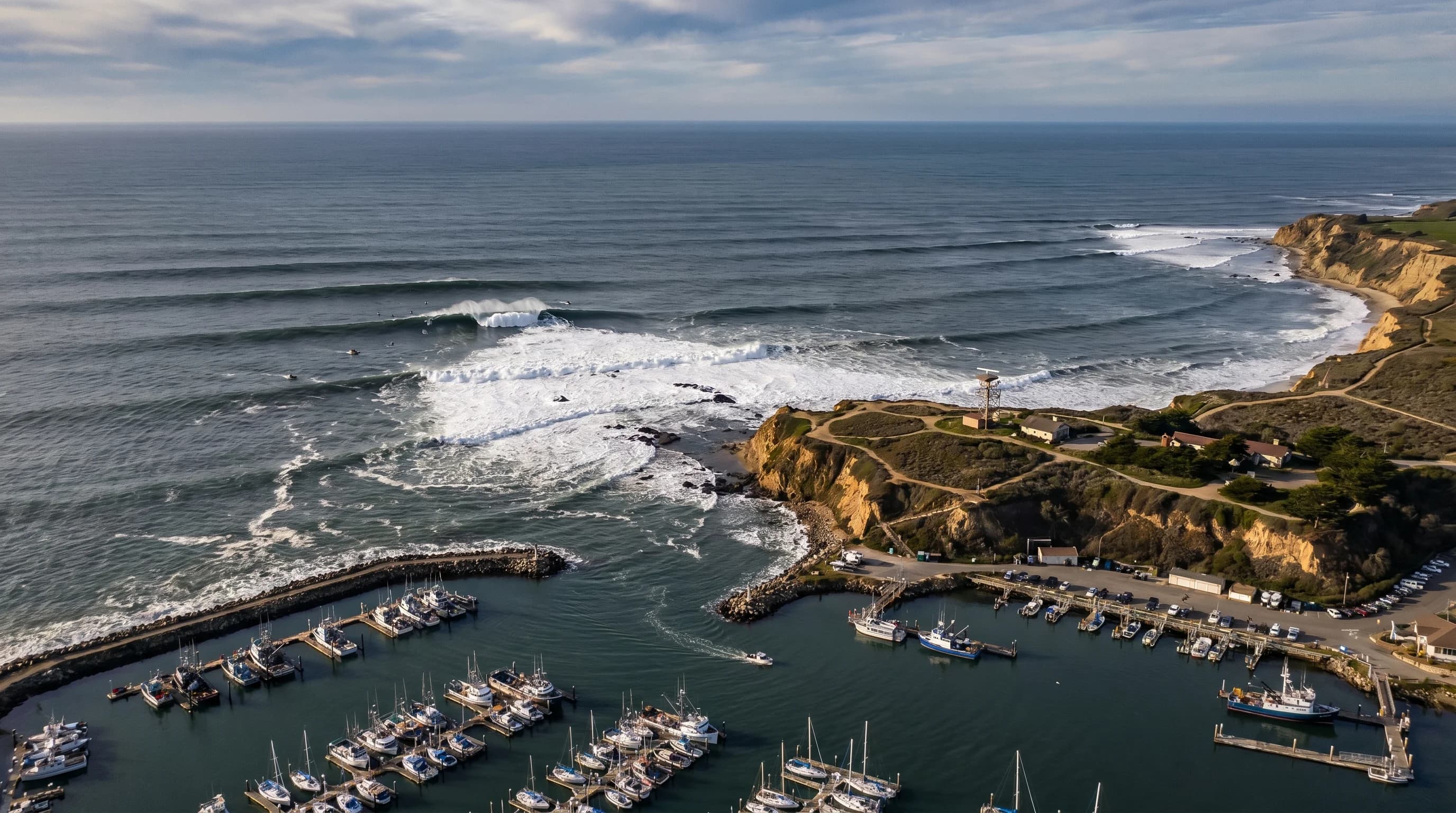 Mavericks big wave breaking at Pillar Point in Half Moon Bay: a massive 40-50 foot wave face with a surfer in the lineup, white water spray, dark gray winter sky, illustrating the scale and power of the wave at peak season