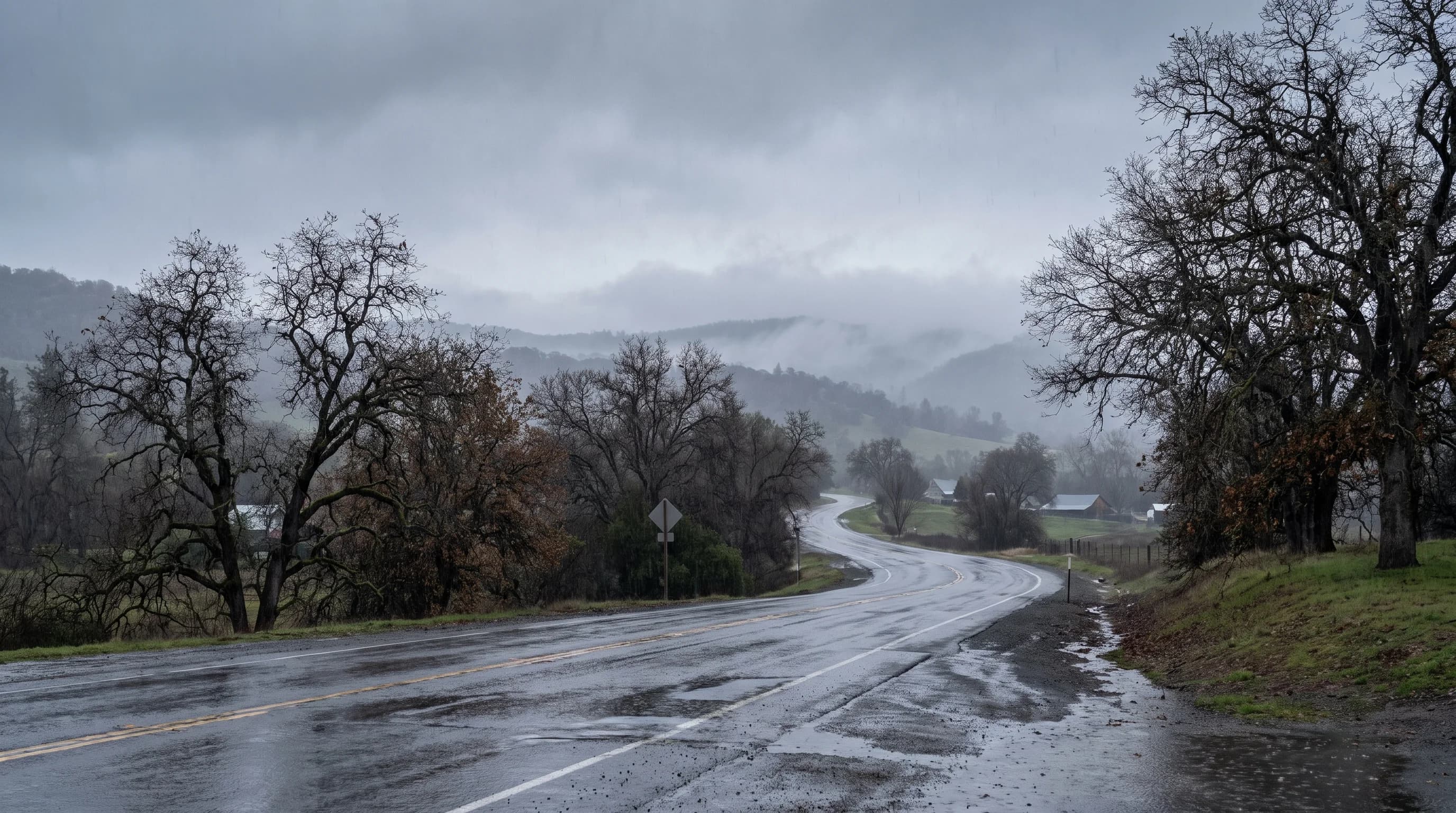 A wet two-lane valley road in light winter rain, its far end softened by mist and low gray cloud